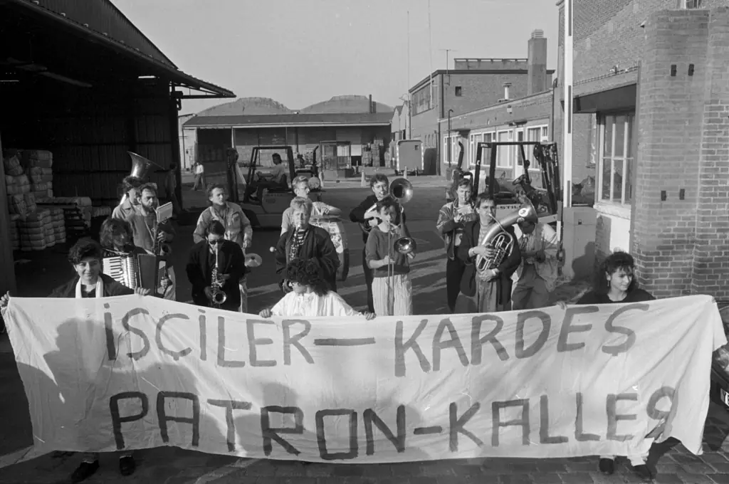 Zwart-witfoto van een groep mensen achter een spandoek met handgeschilderde tekst op het terrein, met blaasinstrumenten en een accordeon; erachter heftrucks, balen en de fabrieksgebouwen