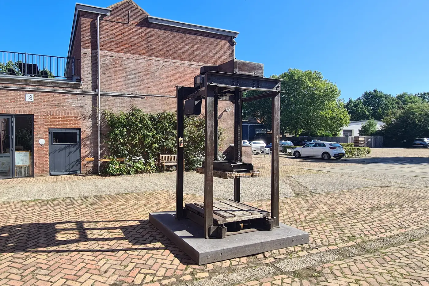 Oude ijzeren balenpers op een lage betonnen sokkel op het klinkerplein, met links de bakstenen gevel met een heg en rechts geparkeerde auto's onder de bomen
