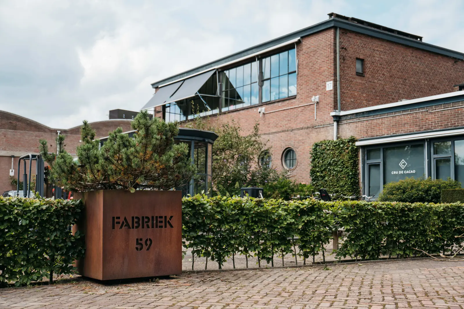 De cortenstalen plantenbak met de uitgesneden letters FABRIEK 59 voor een heg, met daarachter de bakstenen gevel met de glazen serre