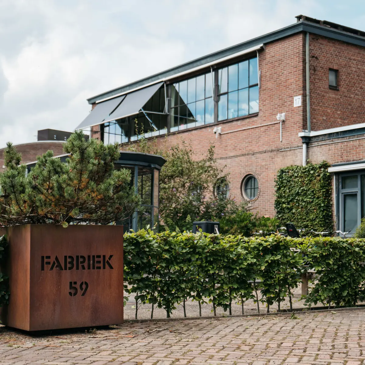 Cortenstalen plantenbak met FABRIEK 59 erin uitgesneden en een den erin, voor een bakstenen bedrijfspand met een beukenhaag en klimop langs de gevel