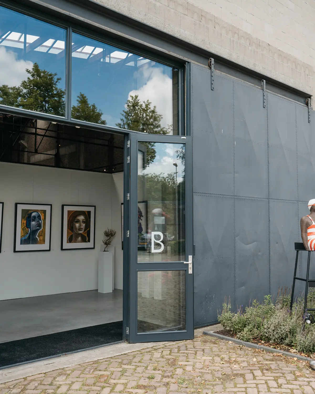 Openstaande glazen deur van een galerie met portretten aan de witte wand binnen, een grijze schuifdeur ernaast en een beeld op een kruk rechts