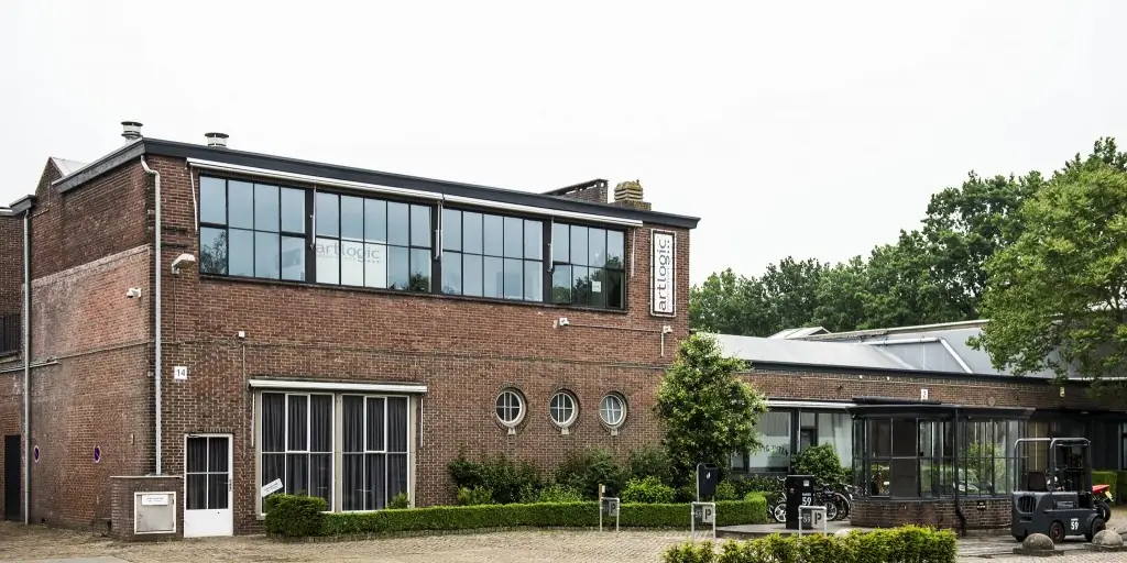 Bakstenen hoofdgebouw met een lange band stalen ramen op de bovenverdieping, drie ronde raampjes eronder en een heftruck rechts op de klinkers