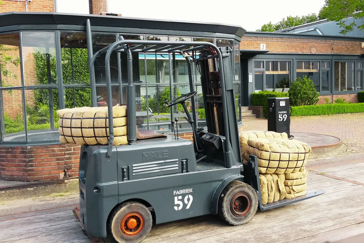 Heftruck met FABRIEK 59 op de zijkant, geladen met balen textiel, voor de glazen serre