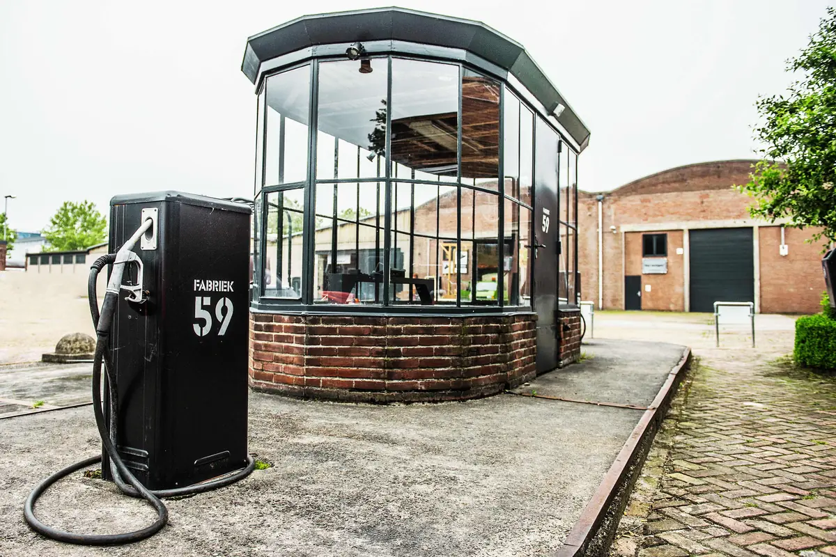 De ronde glazen serre op een bakstenen voet, met een zwarte brandstofpomp met FABRIEK 59 erop ervoor en tongewelfdaken erachter