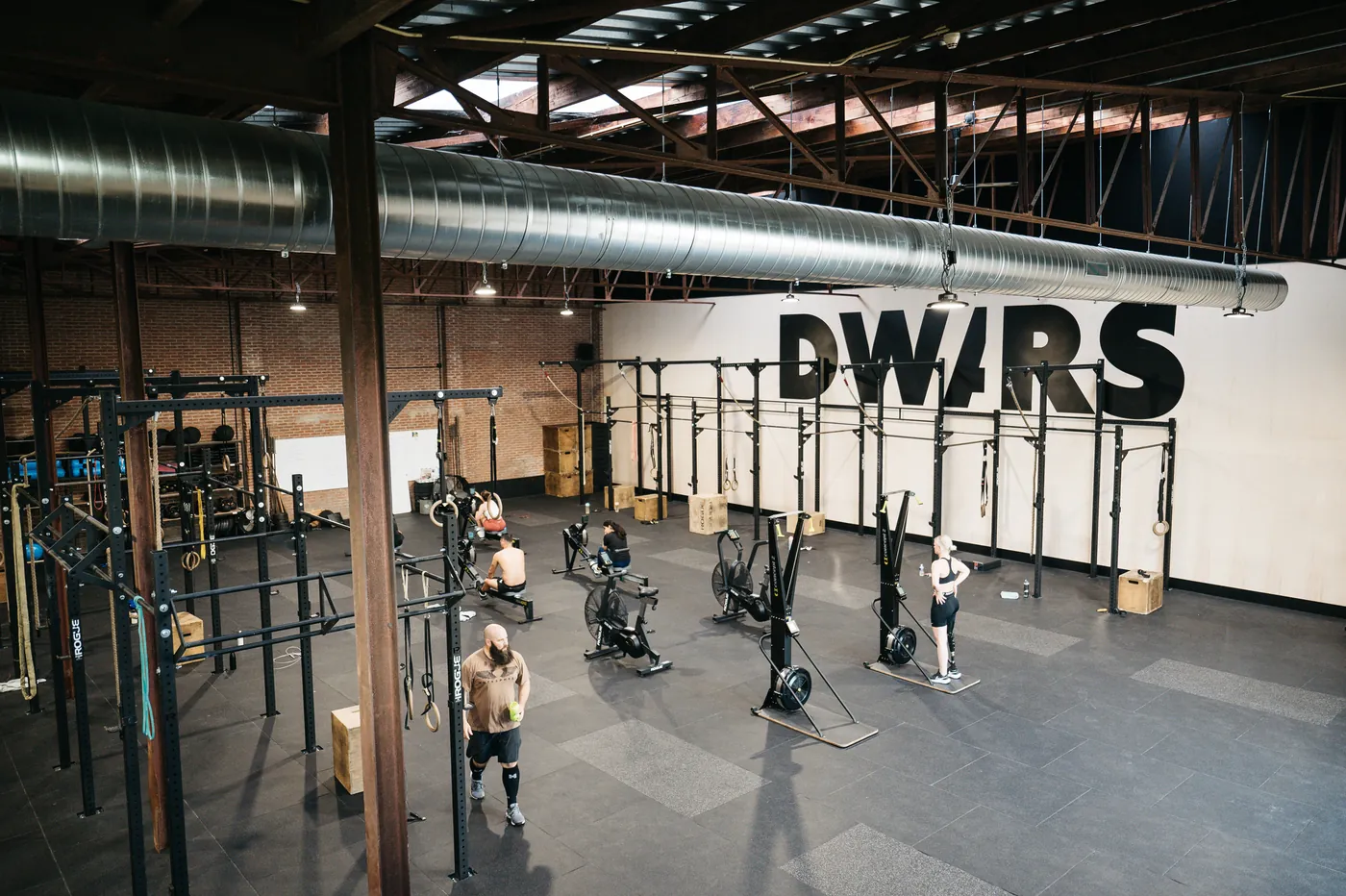 Trainingshal met rekken en roeimachines onder een stalen spantendak, met DWARS groot op de witte wand