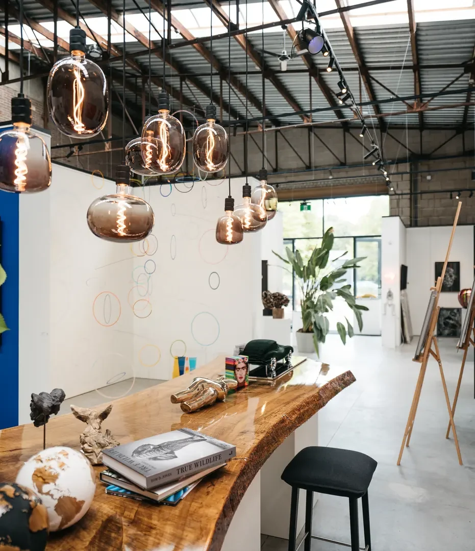Galerie-interieur met koperen hanglampen boven een houten bar, kunstwerken tegen witte wanden en een stalen spantendak