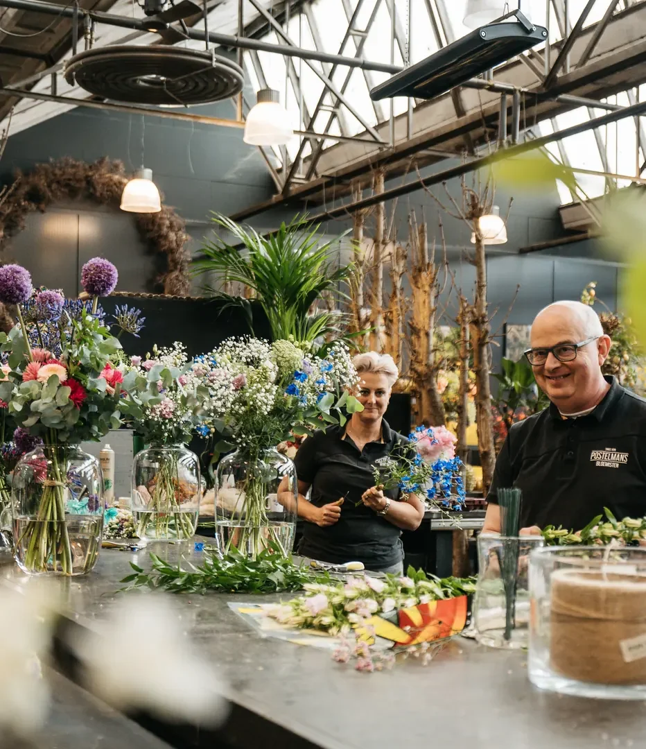 Twee mensen achter een werkbank vol bloemen en groen, onder een zaagtanddak met daglicht
